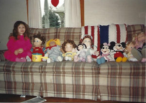 Christine at 3 and a half years old on a plaid couch with a long line of various dolls in front of a blue, white, and red crocheted blanket