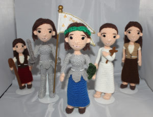 Five crocheted dolls of Joan of Arc in various stages of her life. From left to right: a young shepherdess, a warrior, a saint, a martyr, and a teenage maiden.